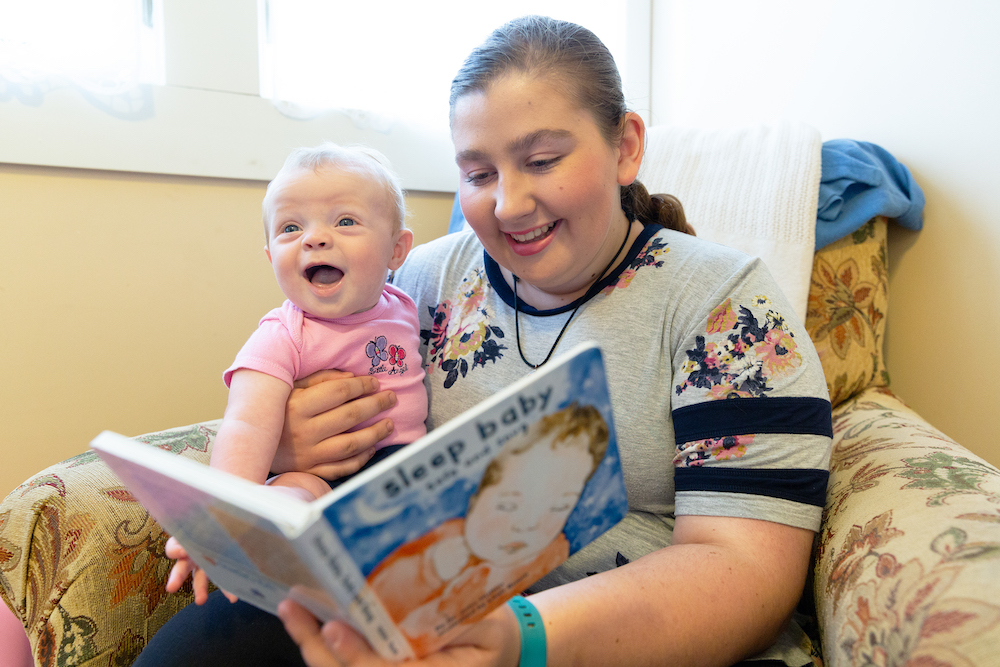 Teenager reads book to smiling baby while they sit on the couch