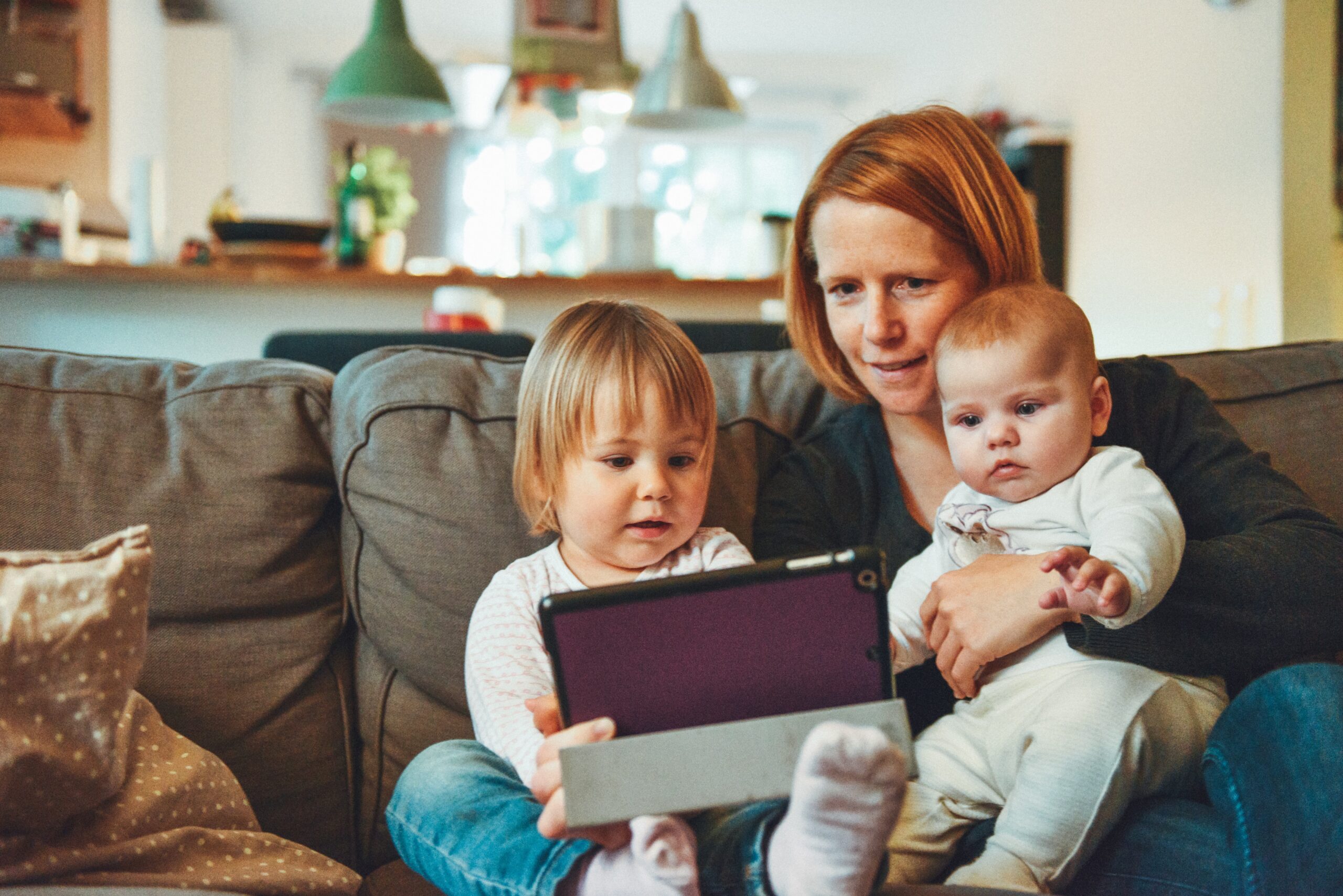 Redheaded mother on couch with toddler and baby looking at ipad