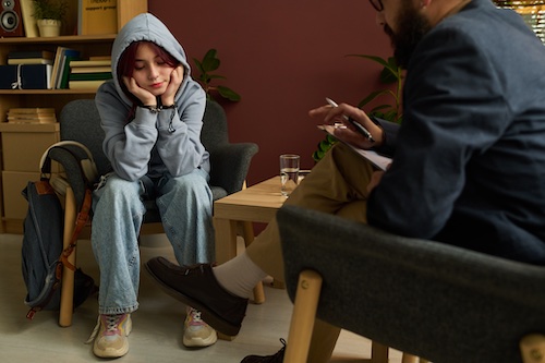Teenage girl in blue casual attire sitting in front of school psychologist and sharing her problems during session