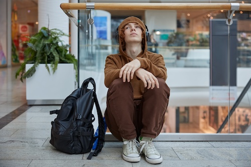 Teenage boy in headphones sitting nearby moving staircase at shopping mall and looking concerned.